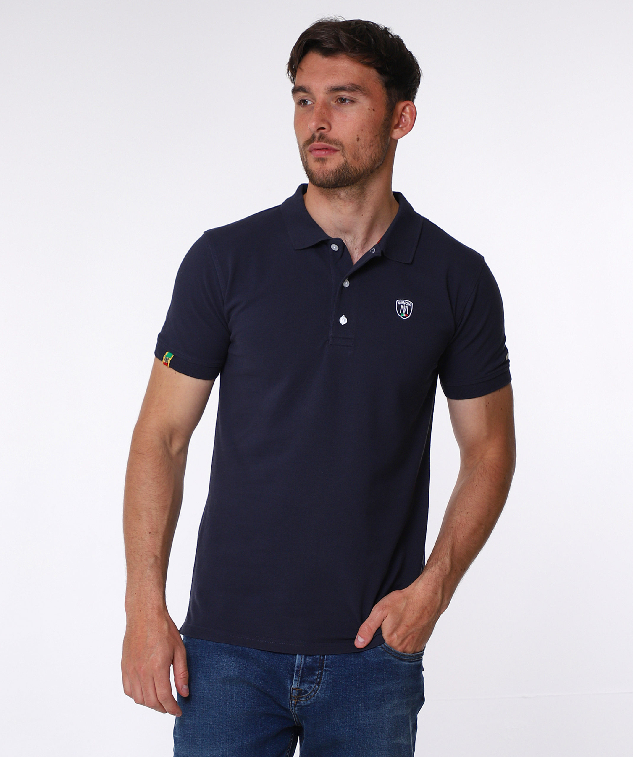 MANCINI - Geoffrey navy polo shirt Navy blue pique cotton polo shirt, with small shield MANCINI logo on the chest and a small logo on the left arm. Contrasting black button hole stitching. The word MANCINI under the collar in black print. Red ribbon detail on the bottom inch of the t shirt on both sides.
